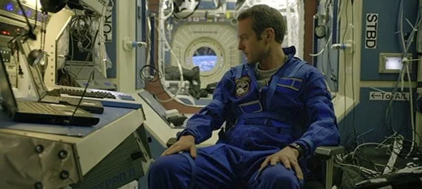 O protagonista sentado olhando os computadores da estação espacial em órbita de um planeta Terra que ficou silencioso de repente.